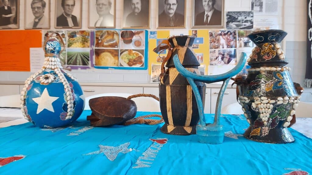 Three large, handmade sculptures on a bright blue tablecloth, set against a wall of portraits and posters in a classroom or gallery.
