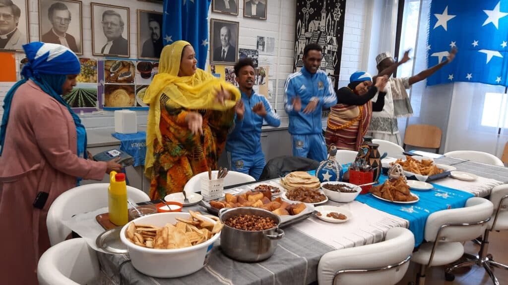 Group of people in colorful traditional dress dancing beside a long table of snacks in a room with blue party banners and portraits on the wall.