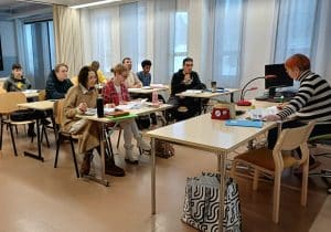Classroom scene with students seated at desks facing a teacher at the right using papers and a computer setup on the desk nearby  Kainuun Opisto